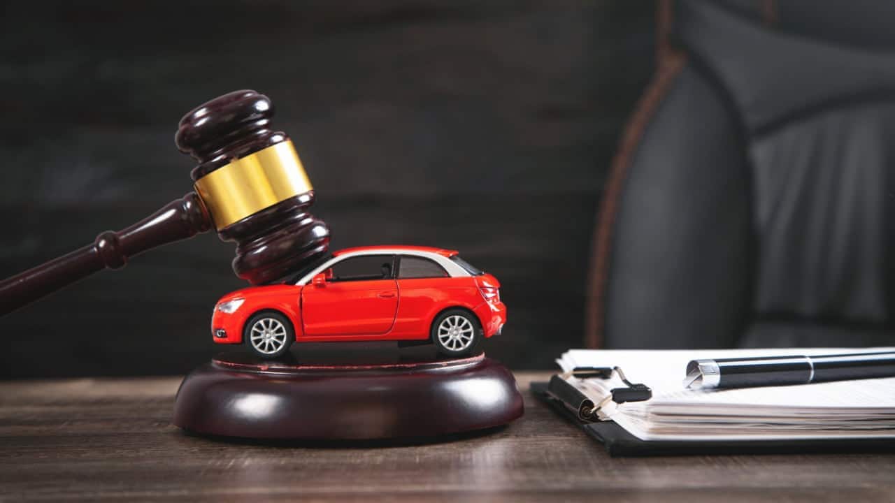 Em cima de uma mesa, malhete de juiz sobre um miniatura de carro na cor vermelha e prancheta com papéis e caneta.
