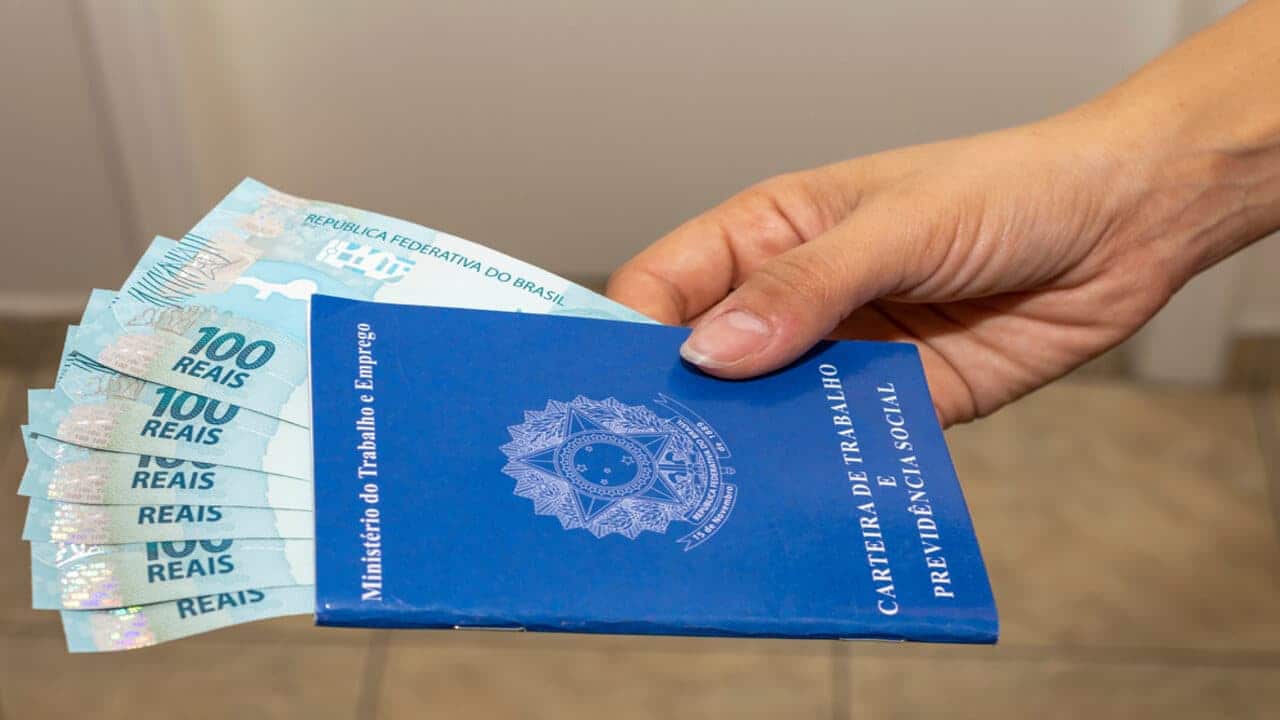 MĂŁo masculina segurando carteira de trabalho com notas de dinheiro no valor de um salário mĂnimo dentro da carteira.