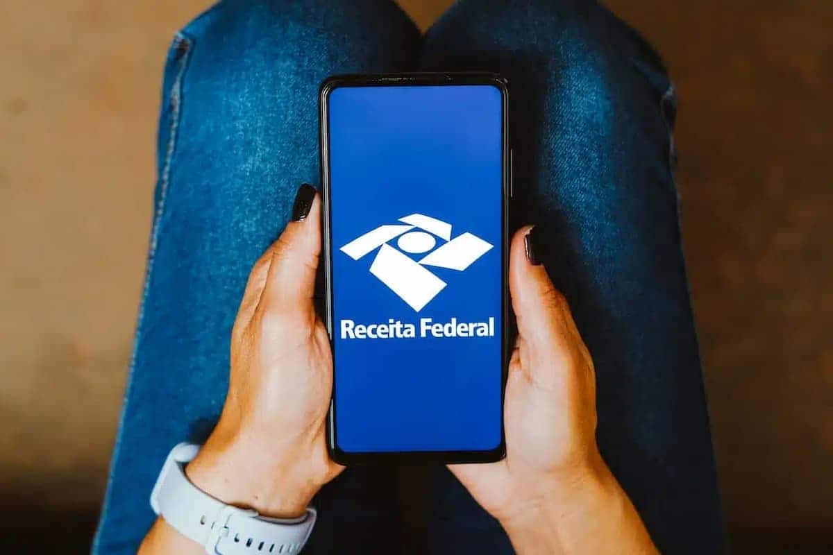 A imagem mostra uma pessoa segurando um celular com aplicativo ou site da Receita Federal aberto. Imposto de Renda