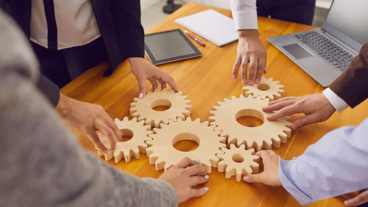 Equipe de trabalhadores de uma empresa juntos, em círculo, posicionando engrenagens de madeira sobre uma mesa de escritório, como metáfora para colaboração, trabalho em equipe, inovação, soluções de tecnologia.