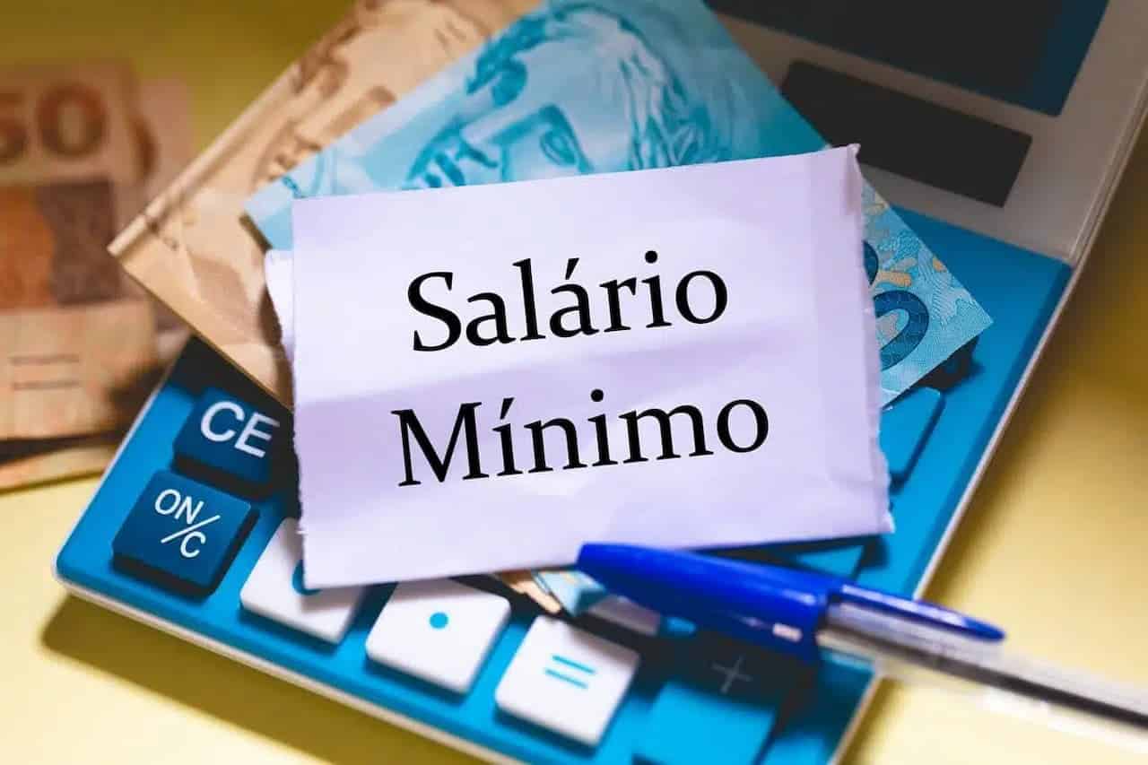 Imagem de uma calculadora com notas de 50 e 100 reais em cima e um bilhete branco escrito "Salário Mínimo". Acima, aparece também uma caneta esferográfica azul.