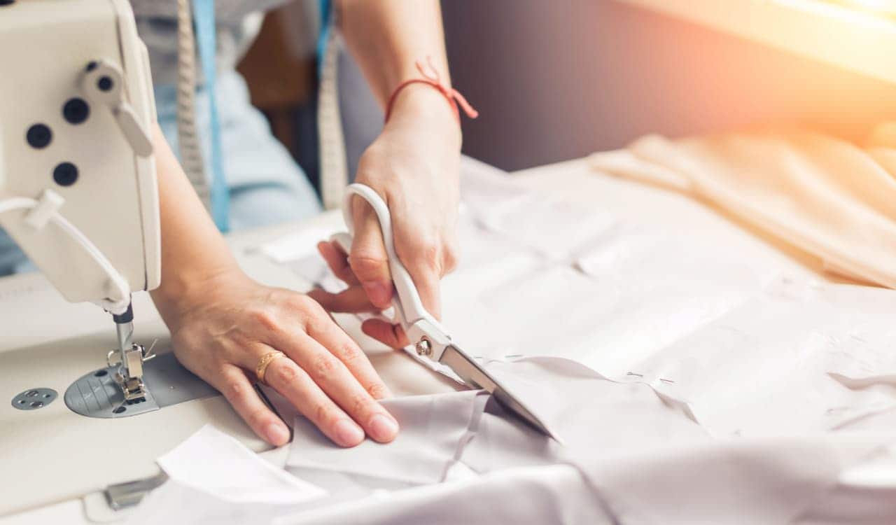 Imagem de mãos de uma mulhar branca que decidiu empreender com costura. As mãos dela estão cortando um tecido branco com uma tesoura sobre uma mesa branca. Ao lado, aparece parte de uma máquina de costura branca.