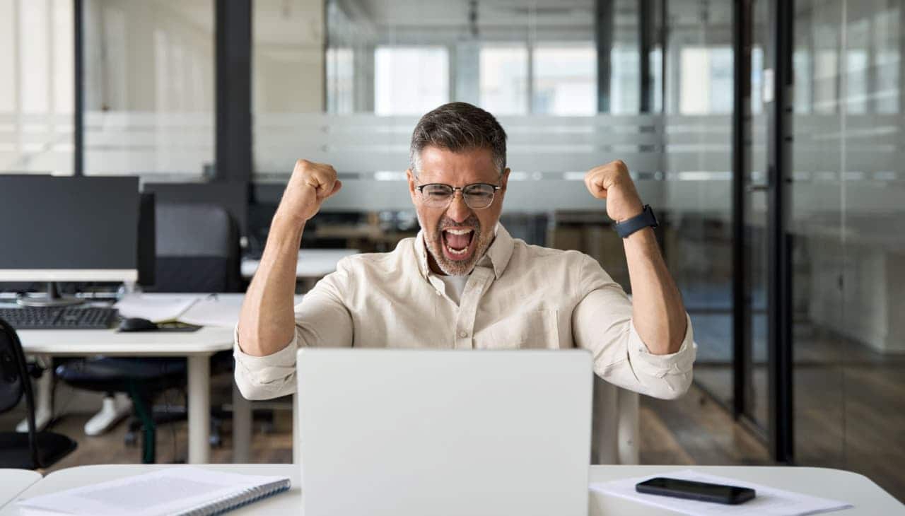 Trabalhador sentado em escritório, olhando para tela de computador, sorrindo e comemorando