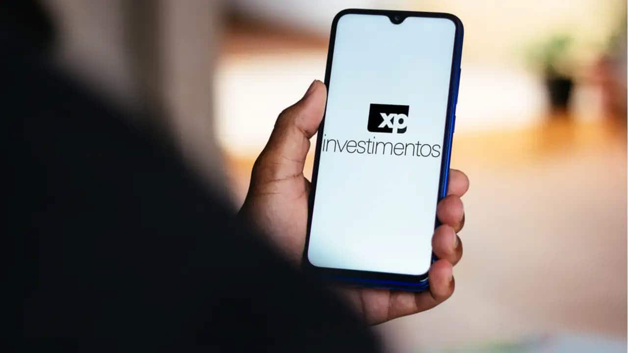 Mão de uma pessoa segurando celular que mostra logo da XP Investimentos
