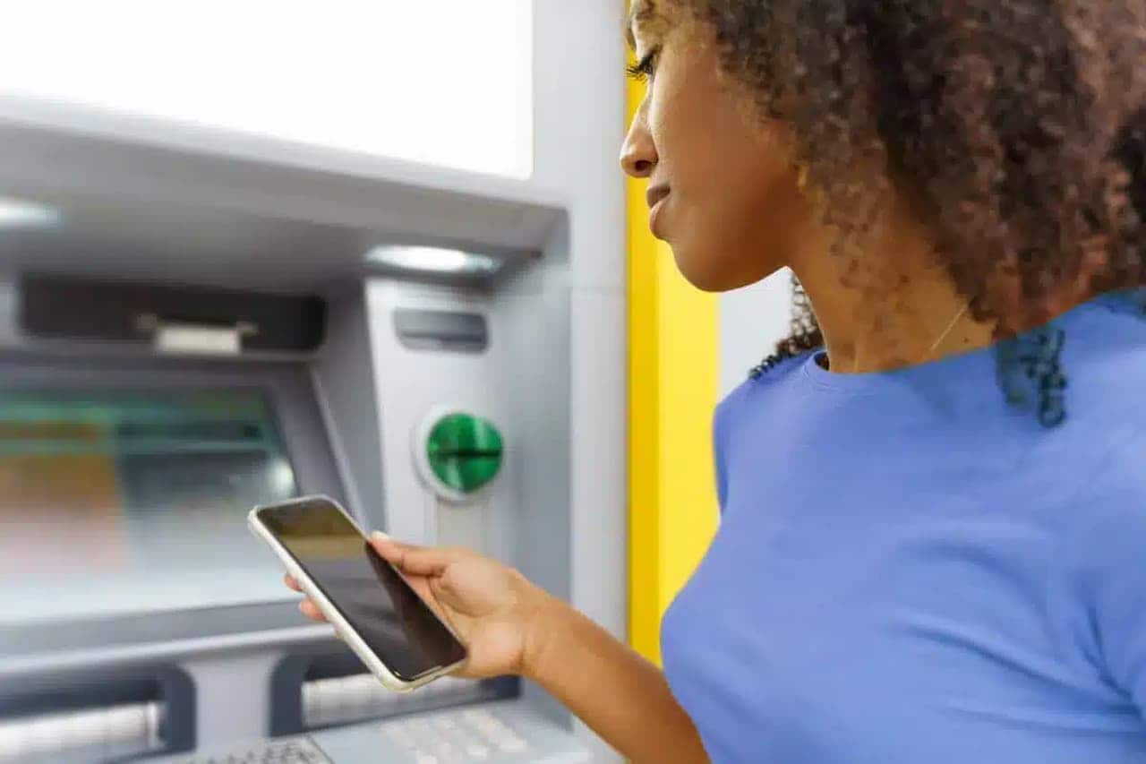 Imagem de mulher mexendo em aplicativo de banco, em frente a caixa eletrônico. DOC e TEC