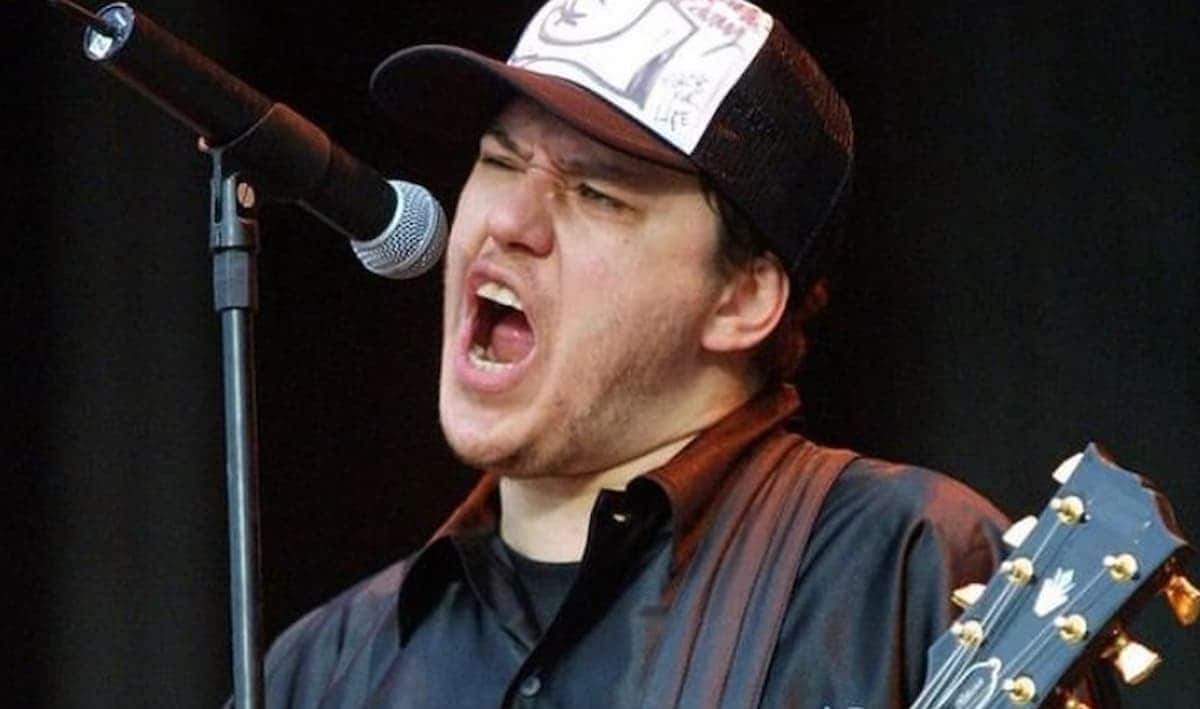 Imagem de Chorão, de boné cantando em uma microfone e uma guitarra