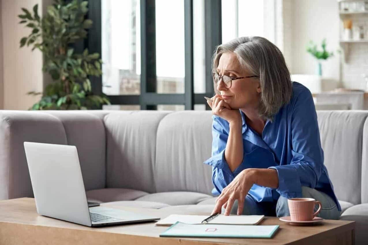 Senhora olhando para um computador aberto em uma mesa, onde há também cadernos e uma xícara. aposentadoria governo