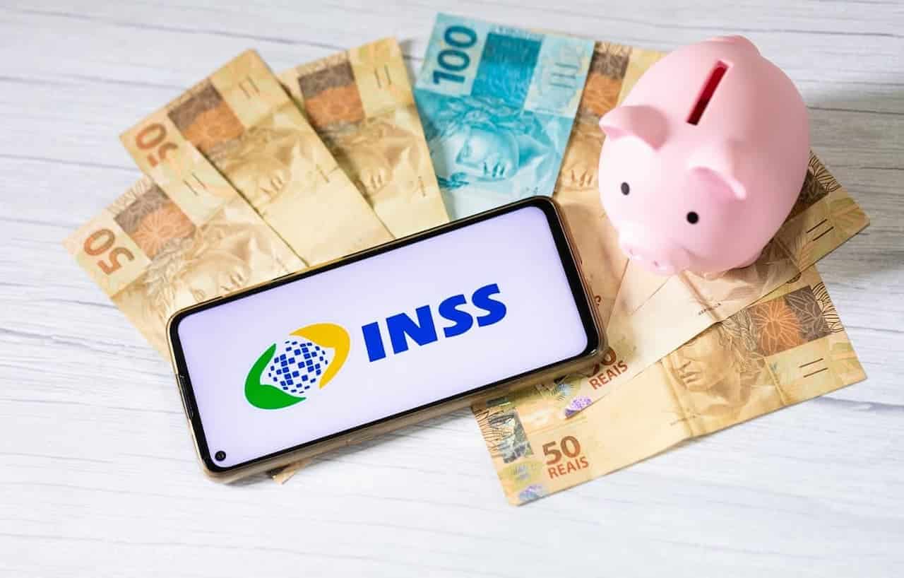 Imagem de um celular com o símbolo do INSS na tela, ao lado, um cofre de porquinho e várias notas de dinheiro INSS
