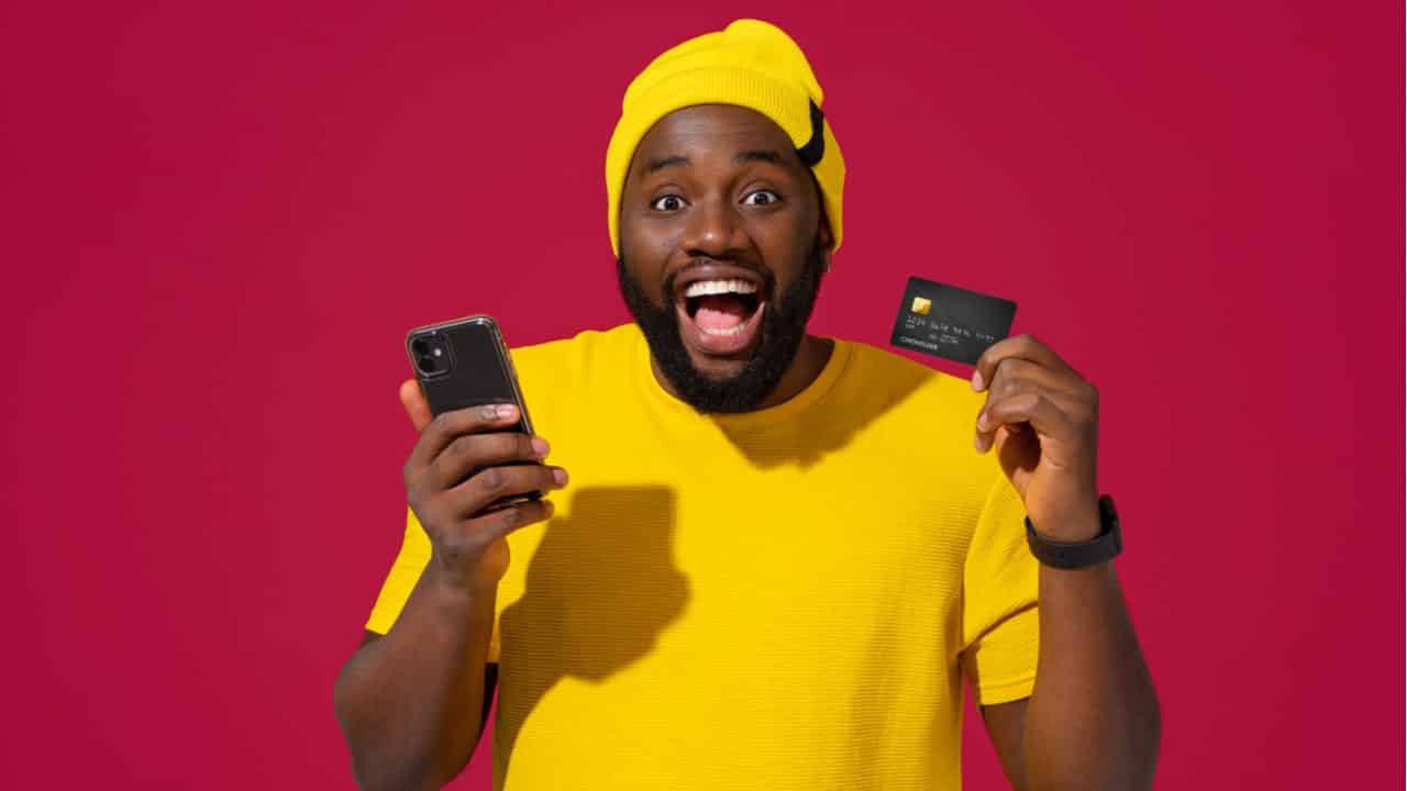 A imagem mostra um homem negro, com roupas amarelas, segurando um cartão Black e celular.