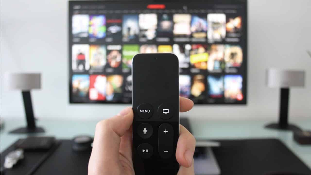 Imagem mostrando mão segurando controle remoto em frente a uma televisão streaming