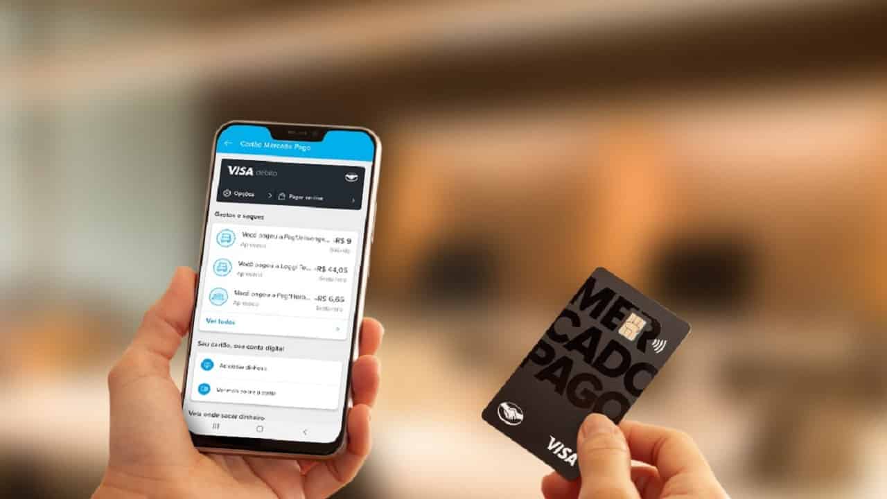 Duas mãos de pessoa branca segurando um celular com o aplicativo do Mercado Pago aberto e um cartão de crédito preto do Mercado Pago