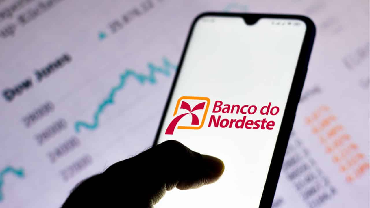 Celular sendo segurado por uma mão e com tela inicial destacando o logo do Banco do Nordeste. Ao fundo dados estatísticos
