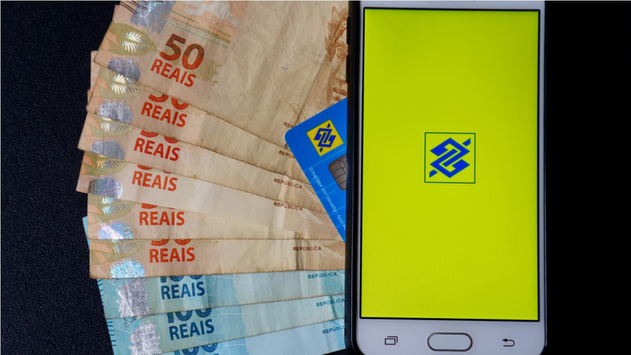 Imagem de um celular com o logotipo azul do Banco do Brasil na tela com um fundo amarelo. Ao lado do celular, aparecem várias cédulas de 100 e 50 reais formando um leque, indicando um empréstimo do Banco do Brasil empréstimo