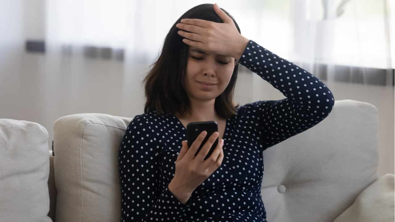 Mulher sentada com a mão na testa e expressão de lamentação enquanto olha para a tela de um celular