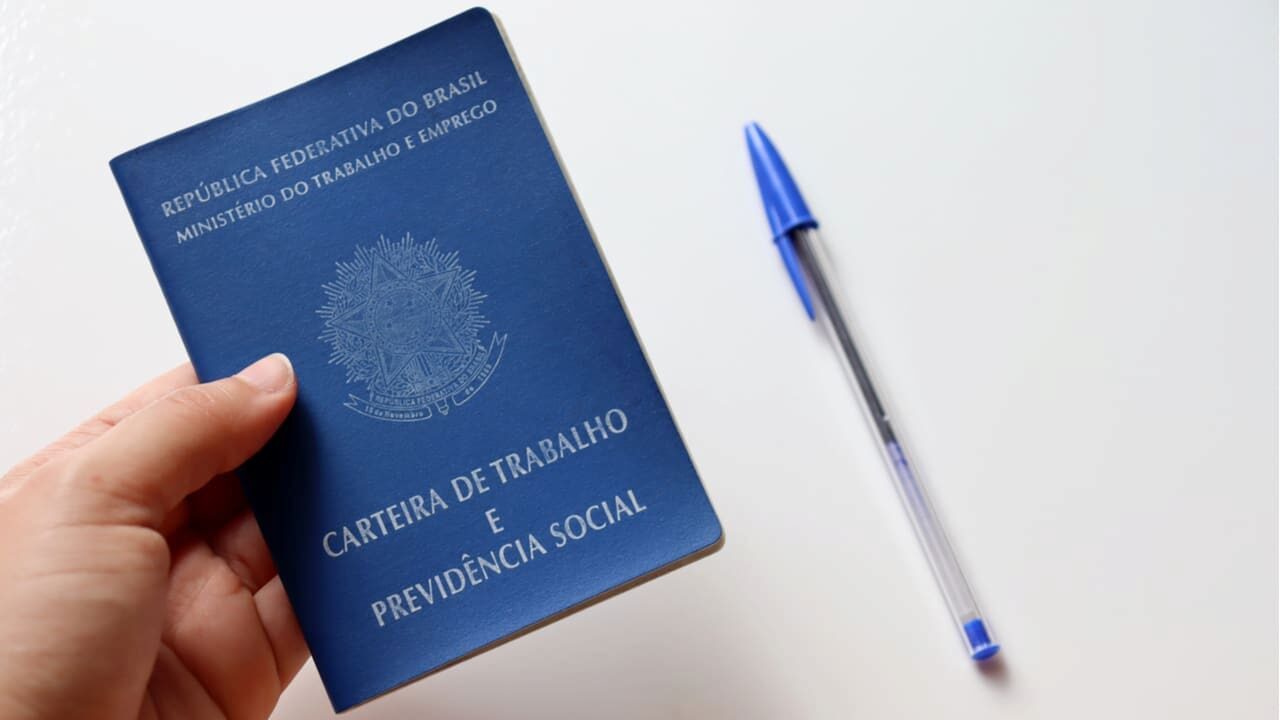 Uma mão segurando a carteira de trabalho e uma caneta azul sobre a mesa. leis trabalhistas
