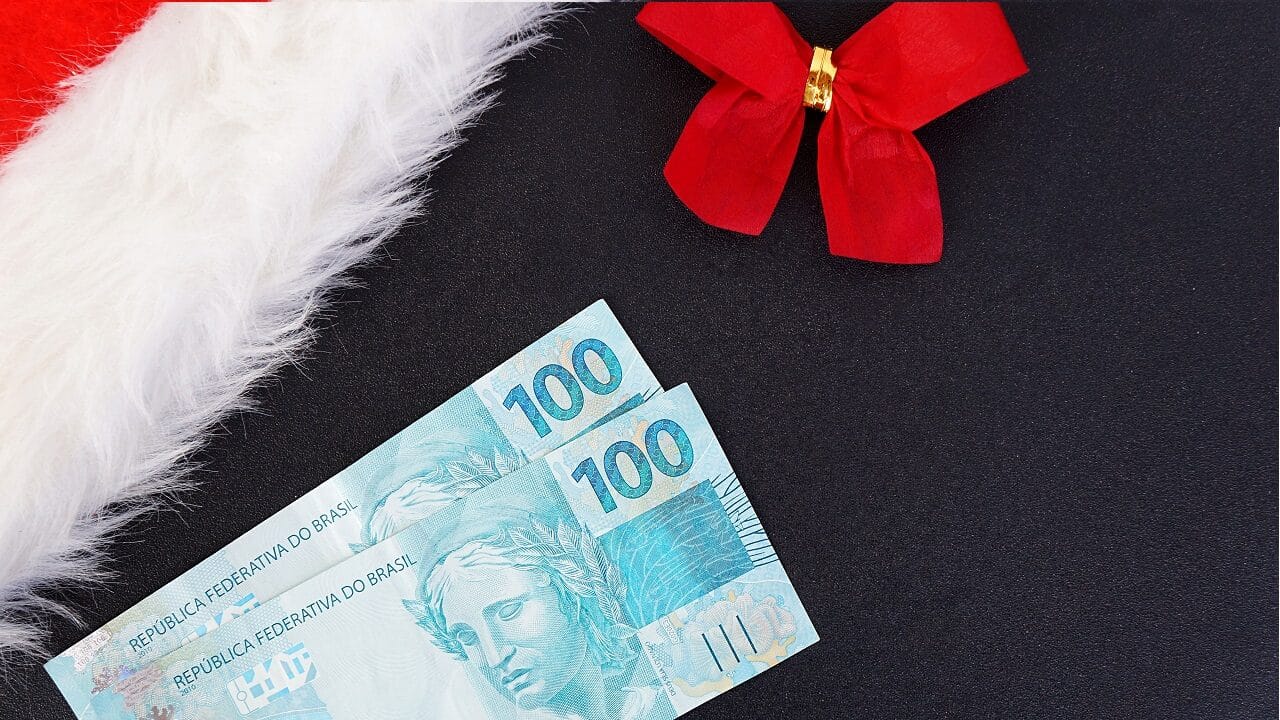 parte de chapéu de Papai Noel com pequeno laço vermelho ao lado e duas notas de 100 reais representando o benefício de Natal e ano novo