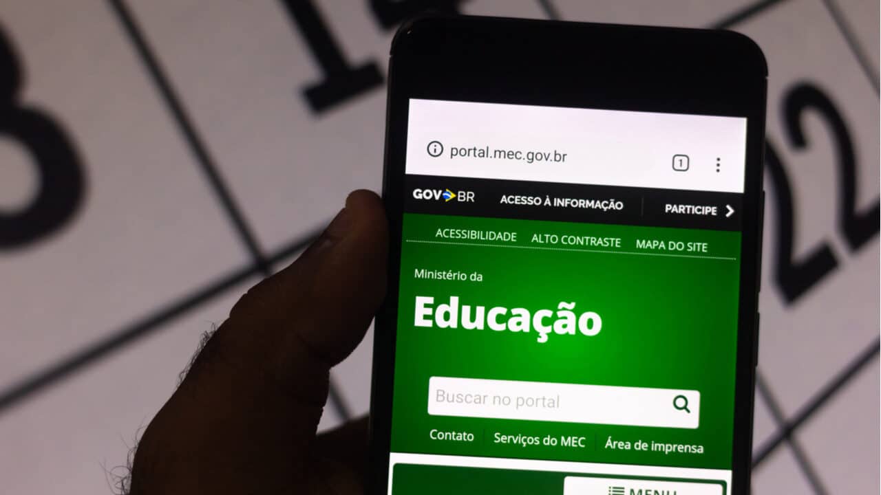Celular mostra página inicial do site do MEC (Ministério da Educação) com calendário ao fundo MEC