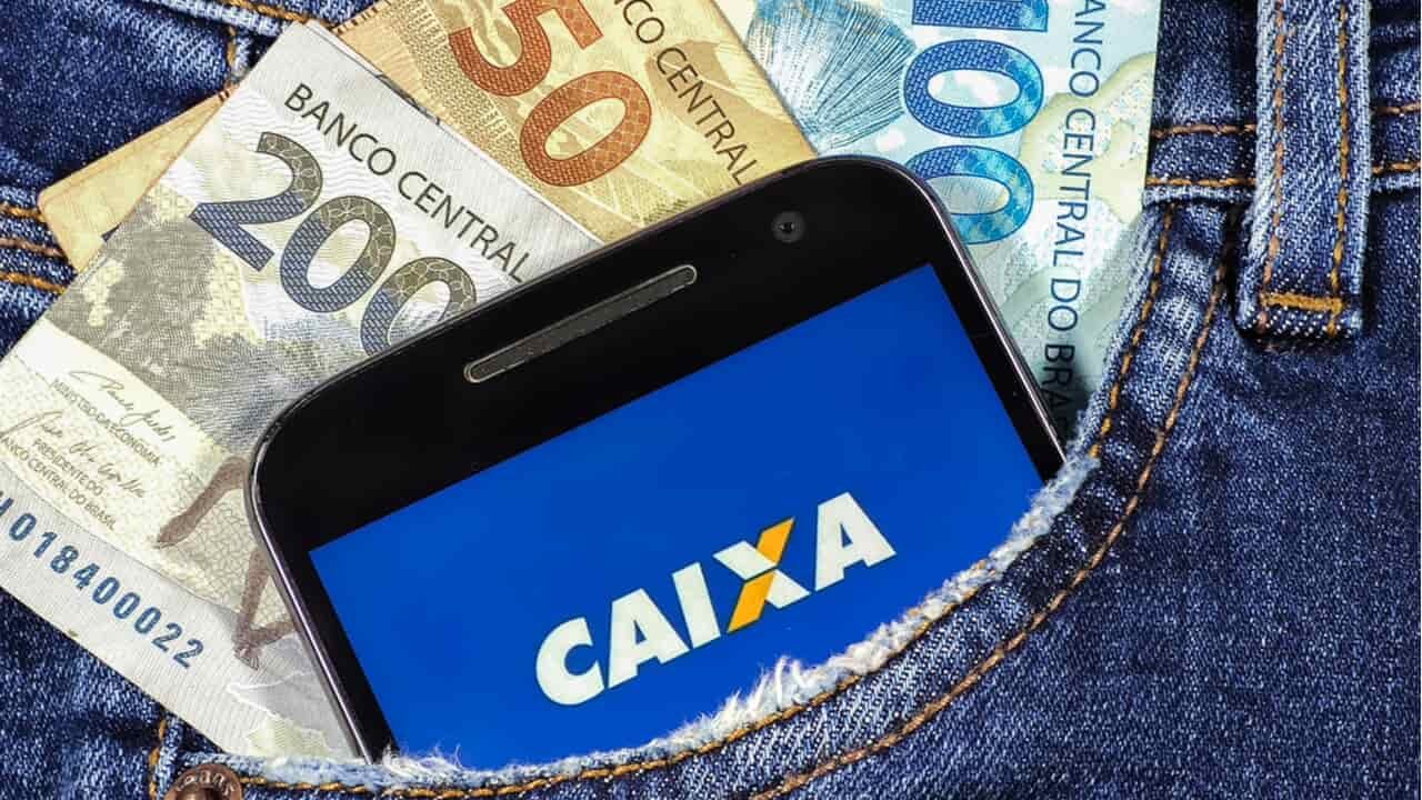 Foto do bolso de uma calça jeans com um celular, mostrando o logo da Caixa, e algumas cédulas de 50, 100 e 200 reais