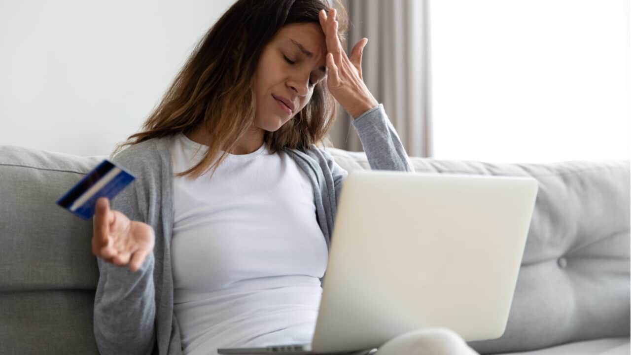 Mulher lamenta com a mão na testa, segurando cartão de crédito e com notebook no colo