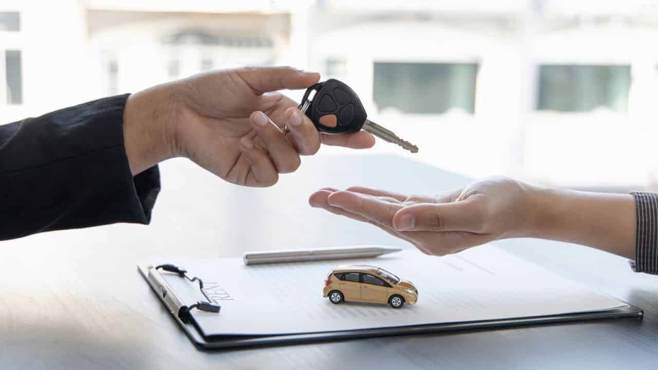 Mão entregando uma chave de carro para outra com um pequeno veículo ao fundo