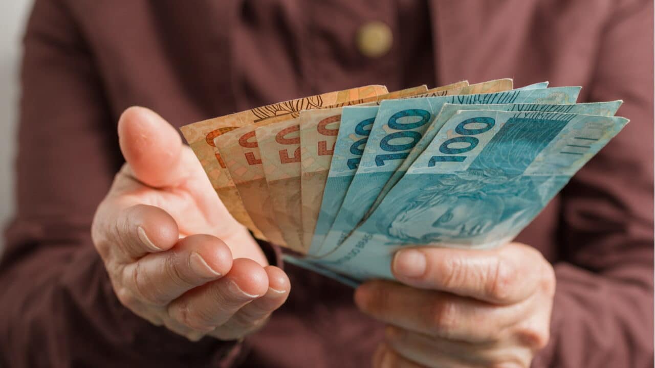 Mãos estendidas segurando notas de 50 e 100 reais