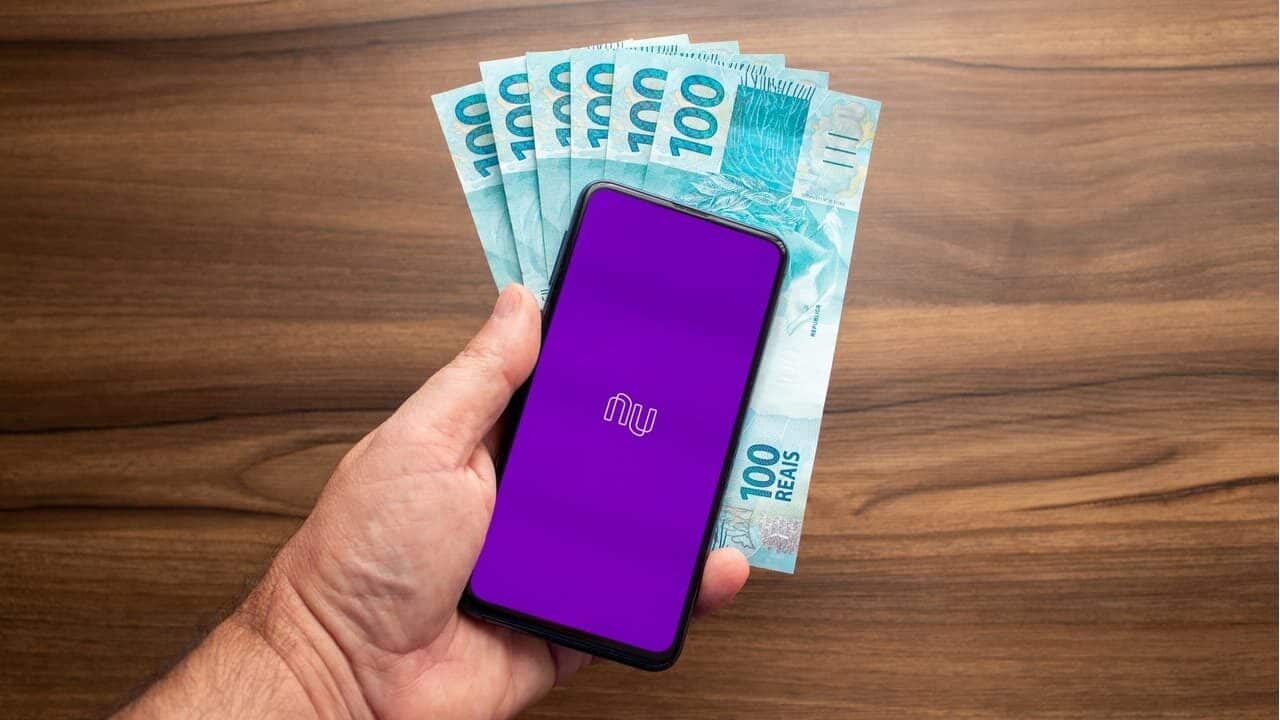 Mão segurando um smarpthone com aplicativo do Nubank e notas de R$ 100 ao fundo