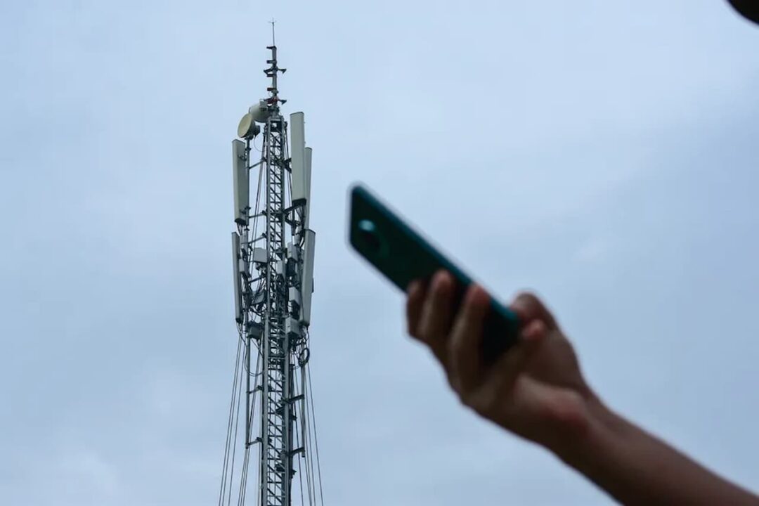 Mão desfocada com celular em primeiro plano, ao fundo da imagem grande antena em evidência
