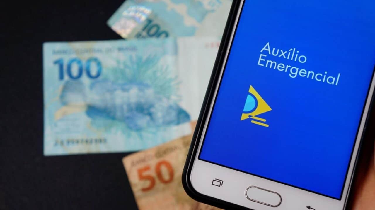 Celular com aplicativo do Auxílio Emergencial aberto e notas de dinheiro embaixo do aparelho