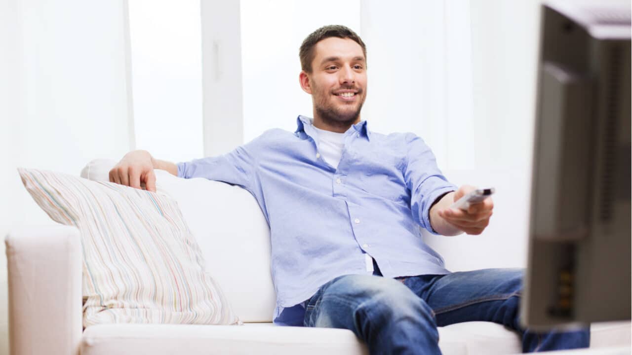 Homem sorridente sentado em sofá e apontando controle remoto para a televisão
