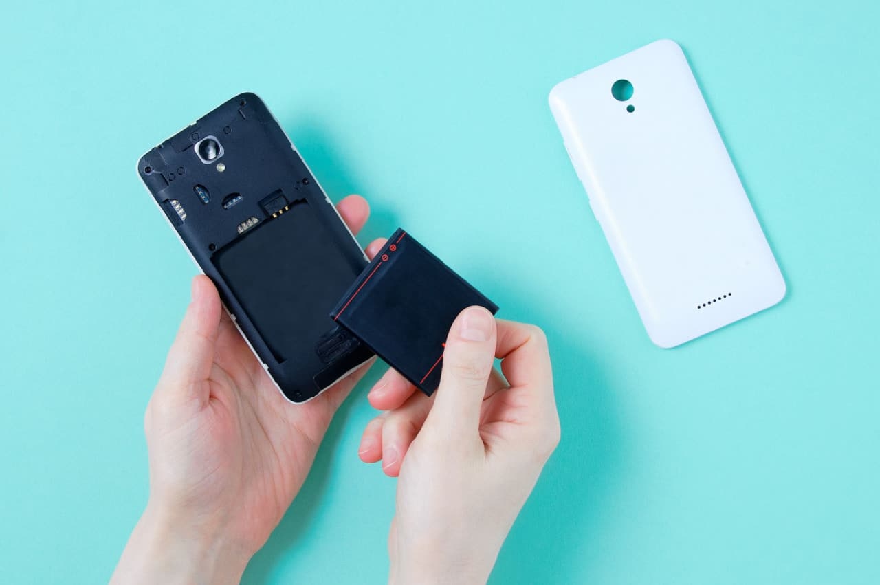 Mãos de uma pessoa retirando bateria removível de celular. Ao lado, a capa traseira do aparelho.