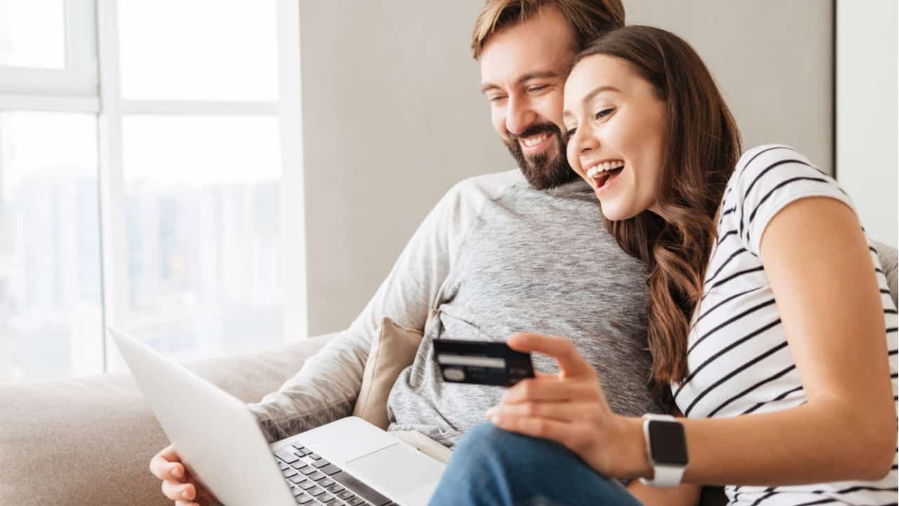 Um casal feliz sentado no sofá com o notebook no colo e um cartão de crédito nas mãos