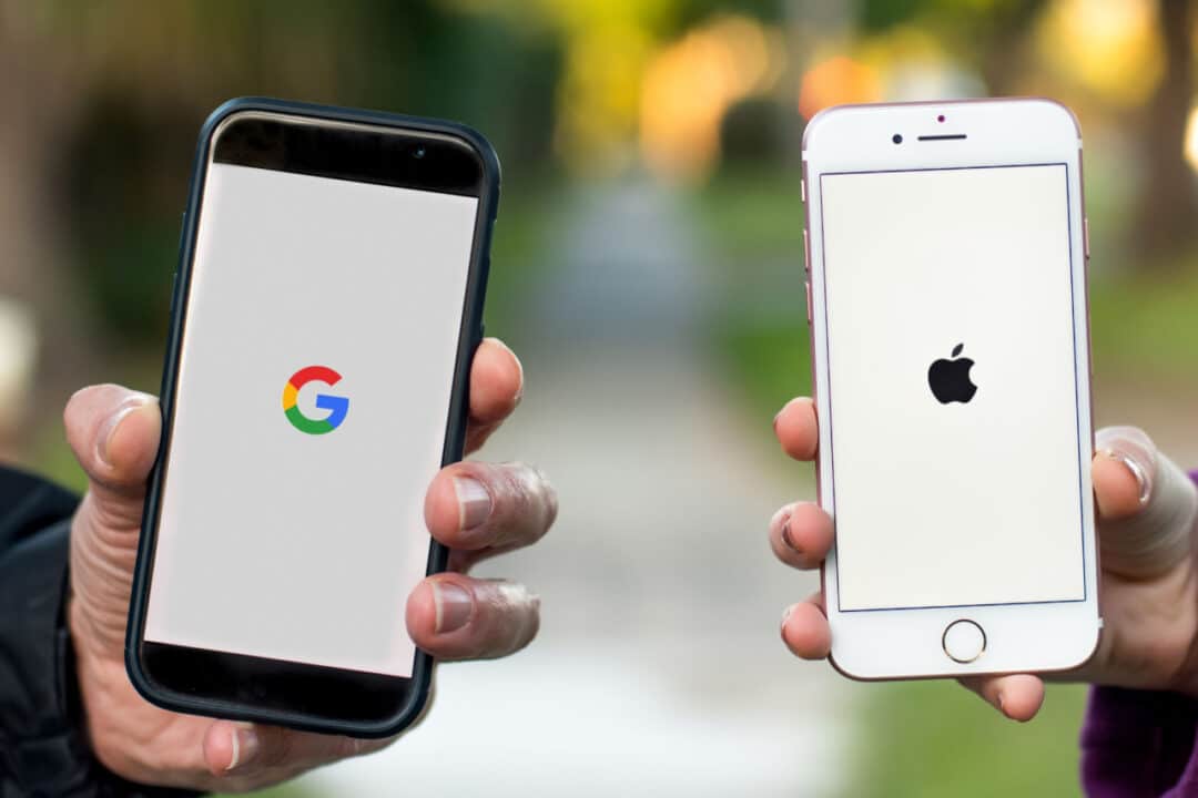 Mão segurando celular Android que mostra logo do Google e outra mão segurando iPhone que mostra logo da Apple