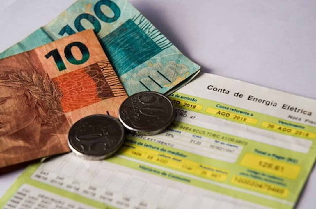 Conta de energia elétrica com dinheiro em cima. Na imagem: uma nota de cem reais, uma de dez reais e duas moedas de cinquenta centavos.