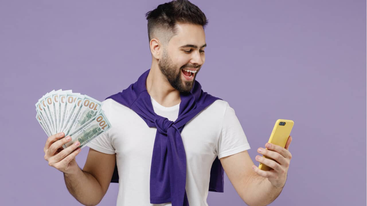 Homem segura dinheiro com uma mão e celular com a outra, sorrindo para a tela