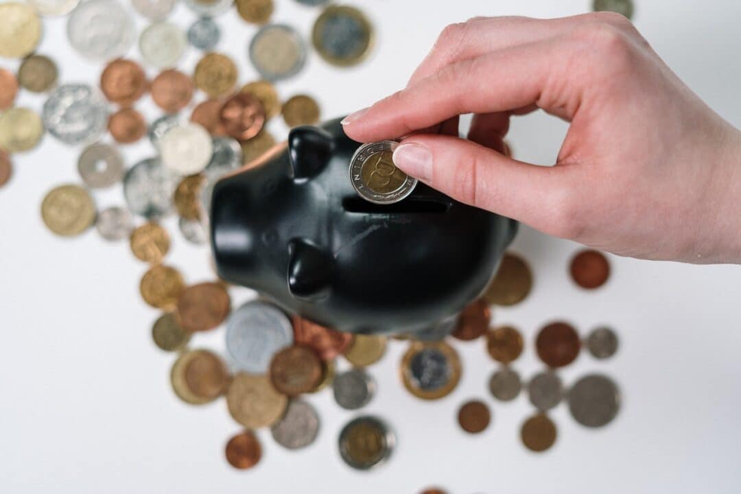 Imagem de uma pessoa guarando uma moeda em um crofre no formato de porco, sendo que existem várias moedas espalhadas na mesa em que o cofre está.