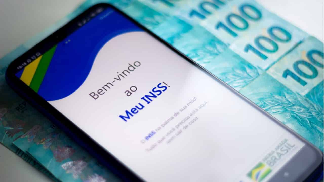 Celular exibindo tela inicial do aplicativo Meu INSS colocado em cima de notas de 100 reais