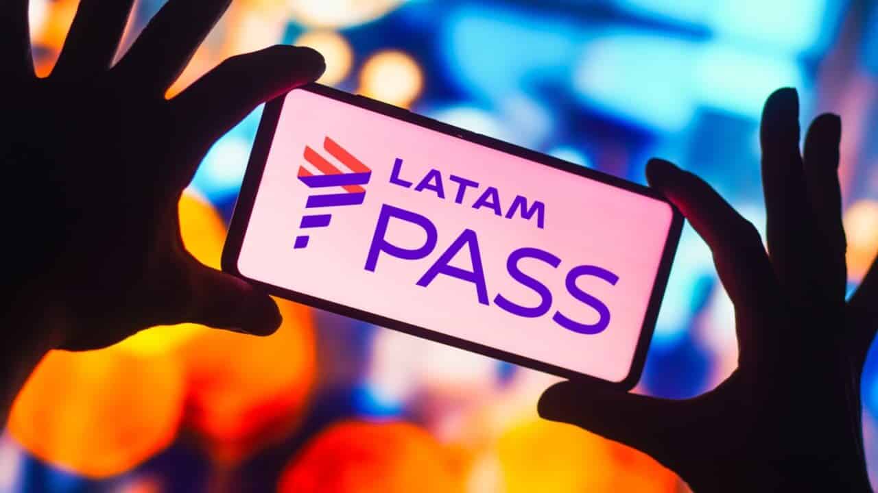 Duas mãos segurando celular que mostra logo do LATAM Pass