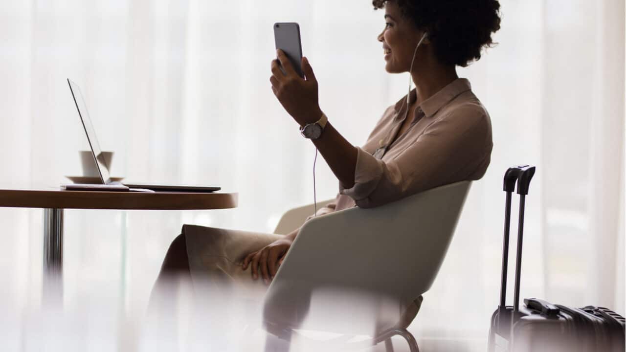 Mulher sentada à mesa em lounge de aeroporto usando celular e fones de ouvido. Sobre a mesa, um notebook aberto. Atrás, uma mala de viagem