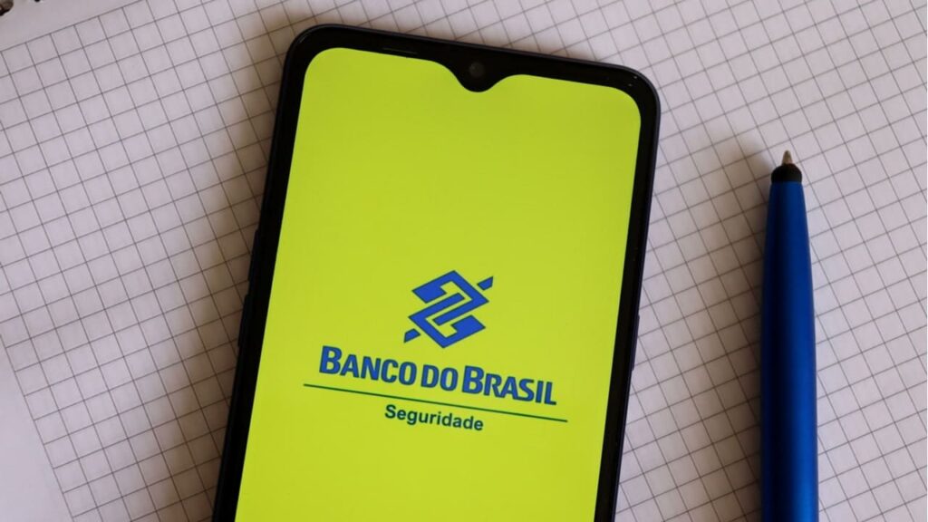 Tela de celular, ao lado de uma caneta, mostrando logo da BB Seguridade ou BB Seguros.