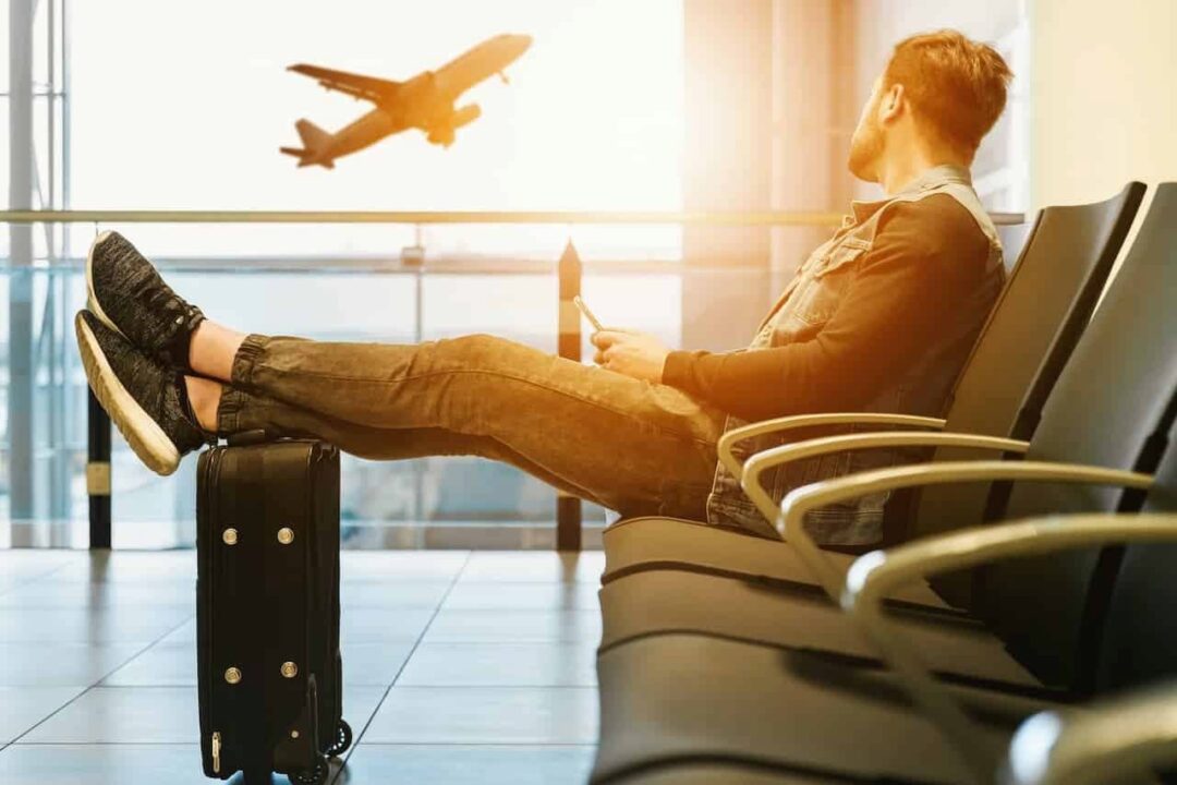 Um homem sentado em uma cadeira dentro do aeroporto olhando para um avião decolando