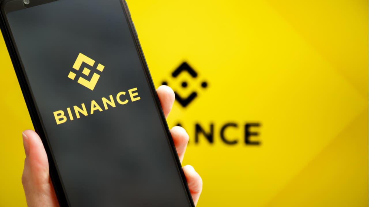 Imagem de um celular com o app da Binance aberto, o logo da Binance se encontra ao fundo
