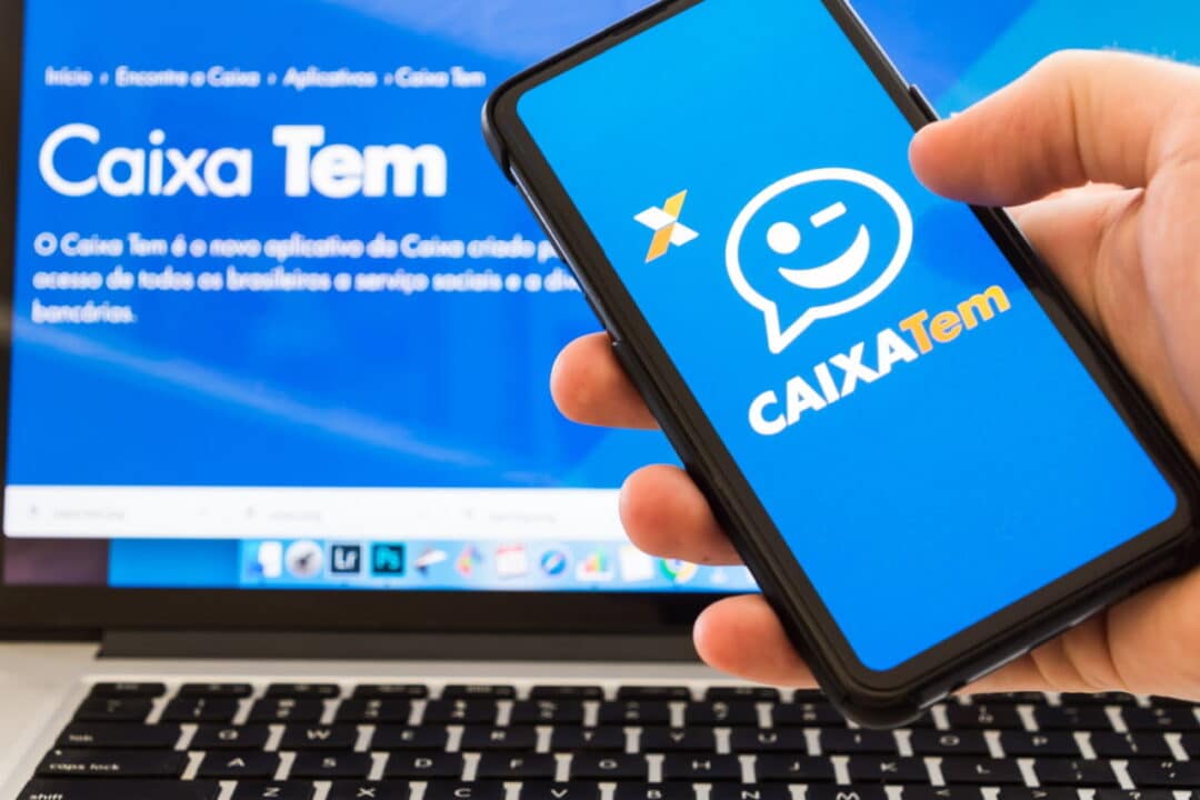 Mão segurando celular que mostra logo do aplicativo Caixa Tem. Ao fundo, tela de computador mostrando página do aplicativo no site da Caixa.