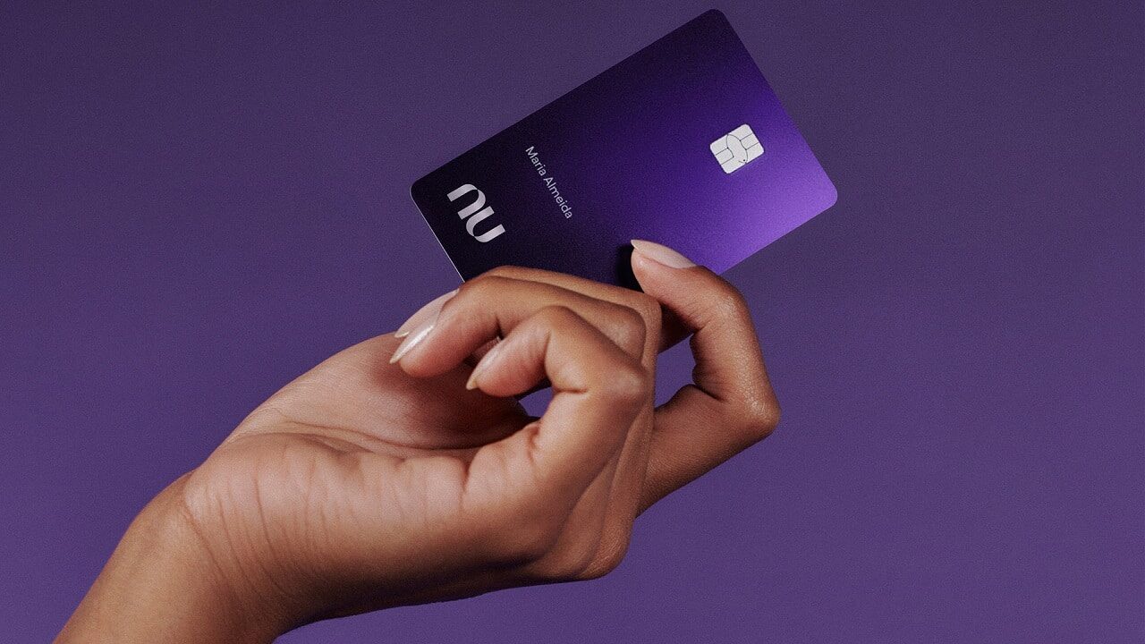 Mão segurando o cartão Ultravioleta do Nubank com fundo roxo atrás