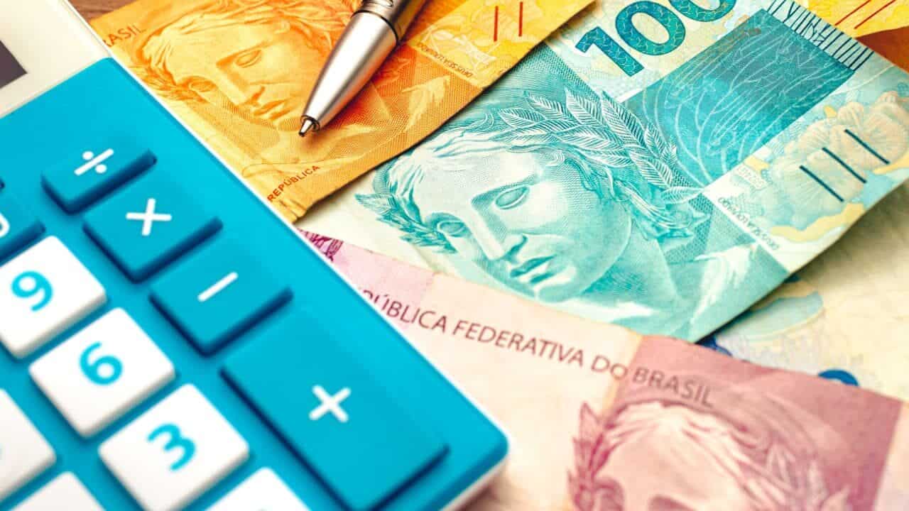 Notas de real brasileiro em uma mesa de madeira com uma calculadora e caneta na composição, representando endividamento.