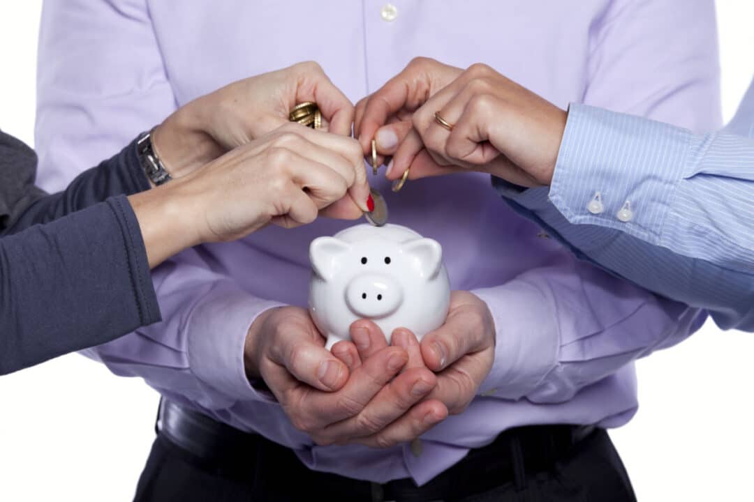 Pessoa segurando cofre em formato de porco com as duas mãos, enquanto outras duas pessoas depositam moedas no interior do objeto representando o consórcio