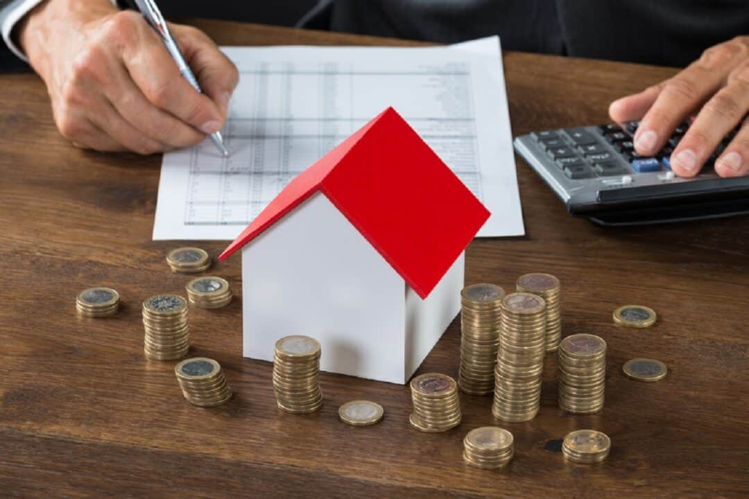 Imagem de miniatura de uma casa cercada por moedas, além de uma pessoa utilizando uma calculadora e um papel.