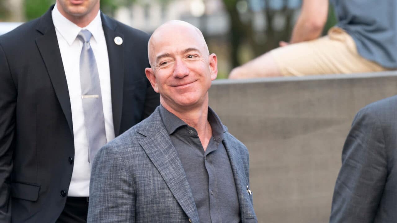 Empresário Jeff Bezos sorrindo em ambiente aberto. Ao fundo, um segurança.