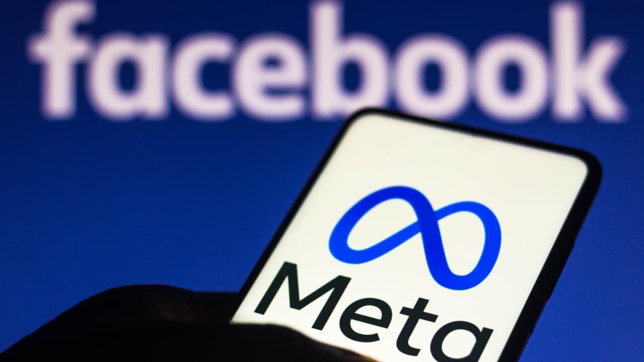 Sombra de uma mão segurando celular que mostra o logo da Meta. Ao fundo, o logo do Facebook.