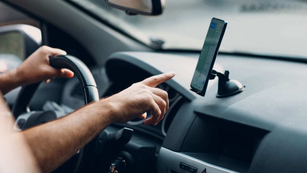 Mão de um motorista, no interior de um veículo, indo em direção a um celular posicionado no painel do carro