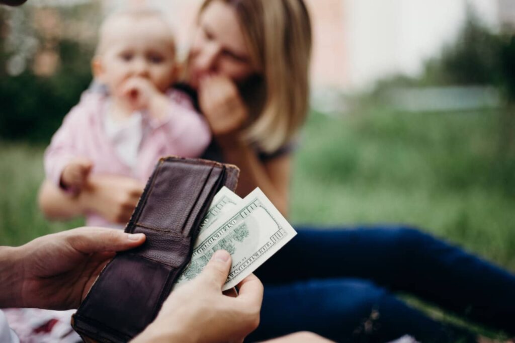 Mãos de uma pessoa segurando carteira e retirando notas de dinheiro. Ao fundo, mãe e bebê aparecem desfocados.